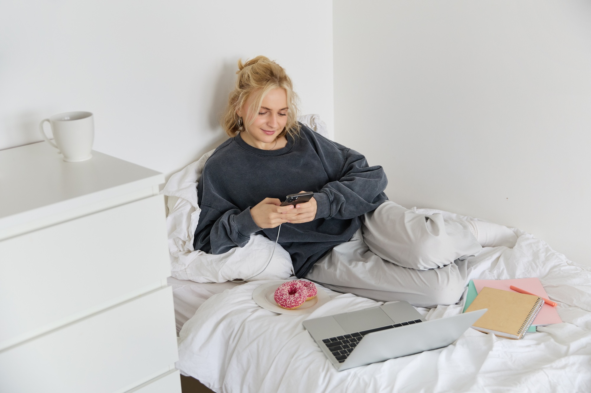 Portrait of smiling candid woman, lying in bed with doughnut, using smartphone and laptop, resting