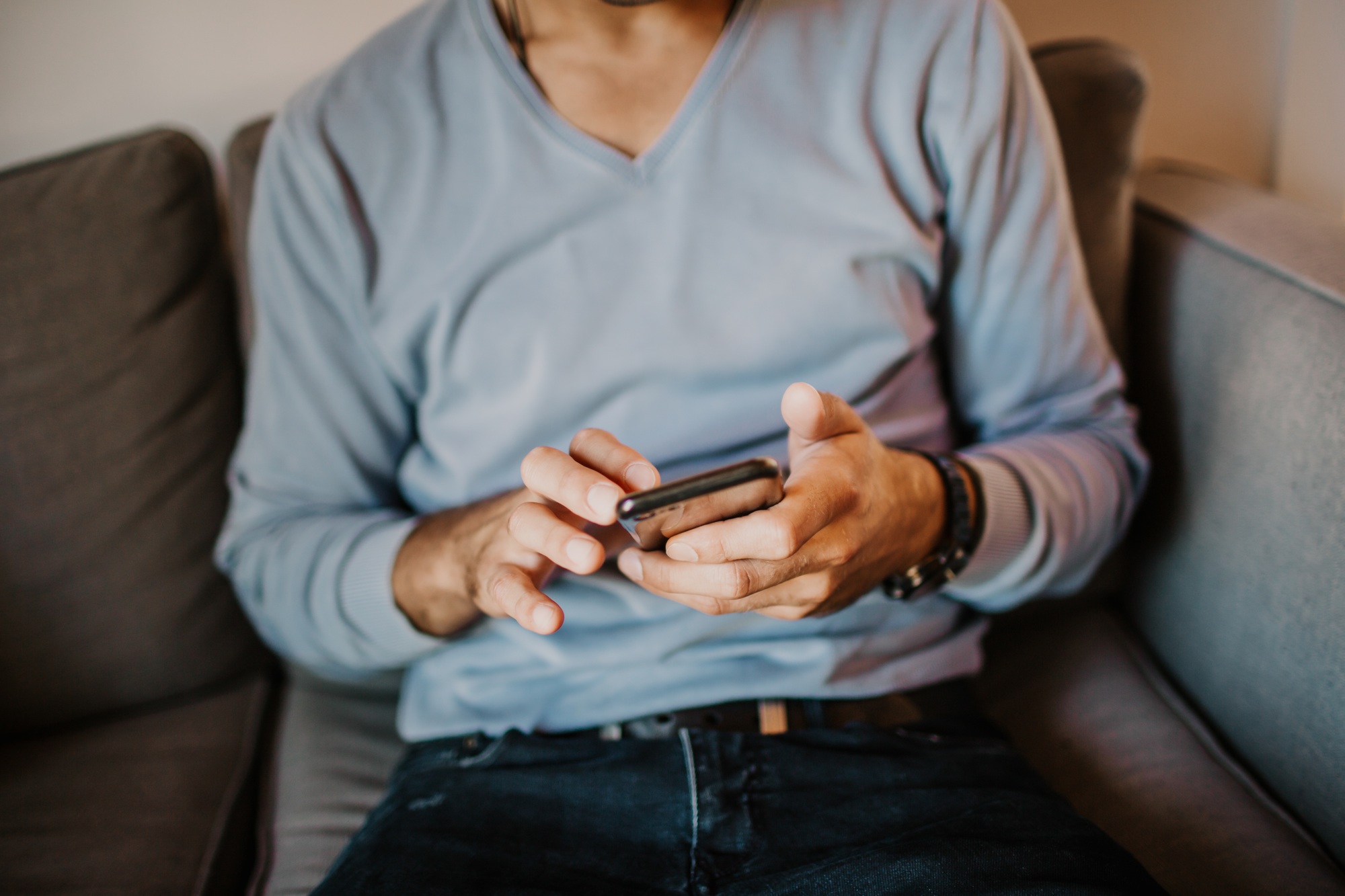 Young handsome man sitting at home on sofa and using mobile phone. Men holding smartphone hands