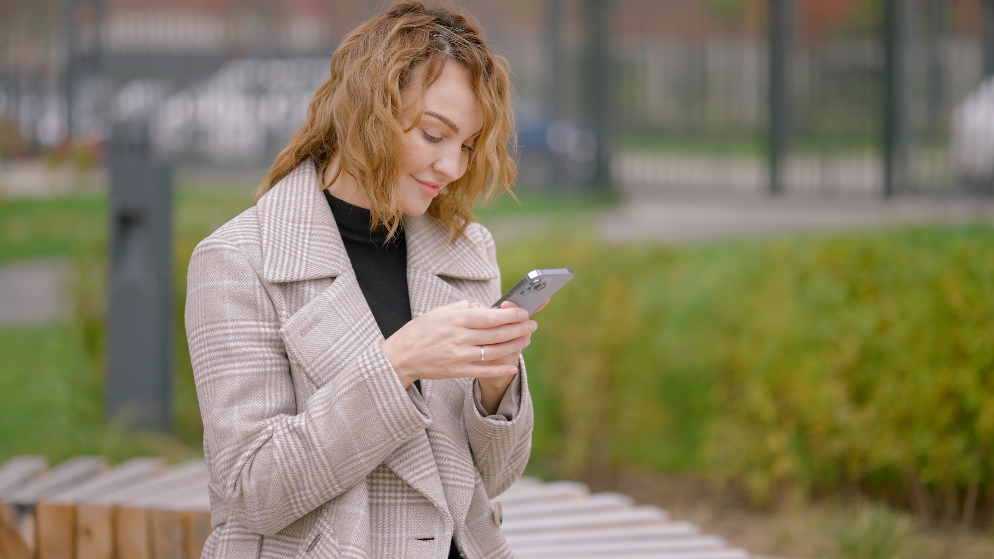 A young woman in checkered jacket looks into a mobile phone while sitting on a bench. Action. Happy