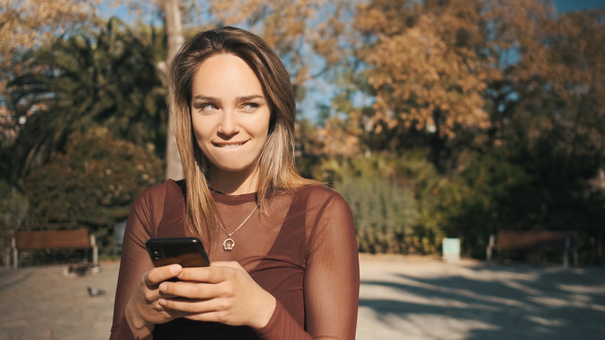 Portrait of beautiful flirty blond girl slyly looking away while texting with boyfriend in park